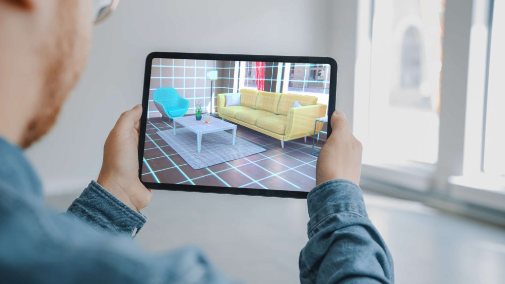 Decorating Apartment: Man Holding Digital Tablet with AR Interior Design Software Chooses 3D Furniture for Home. Man is Choosing Sofa, Table for Living Room. Over Shoulder Screen Shot with 3D Render with augmented reality mobile app